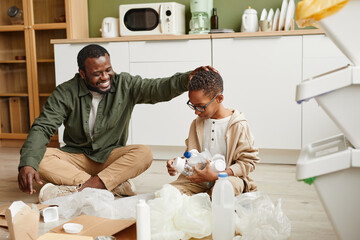 Poster - Full length portrait of happy African-American father and son sorting plastic and paper at home for waste recycling, copy space