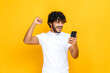 Amazed joyful excited Indian or Arabian guy holds smartphone, get unexpected news, winning lottery, stands on isolated orange background, cheerful facial expression, toothy smile, gesturing with fist