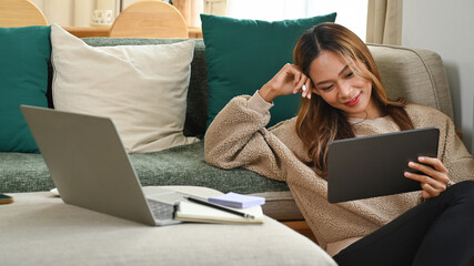 Smiling asian woman watching video on digital tablet in living room.