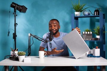 Smiling vlogger holding laptop computer doing online giveaway in vlogging studio with professional live setup. Influencer hosting contest to fans while talking into microphone.