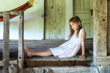 Vintage portrait of a beautiful little girl with bare feet sitting at an open terrace in an old country house with wooden floor