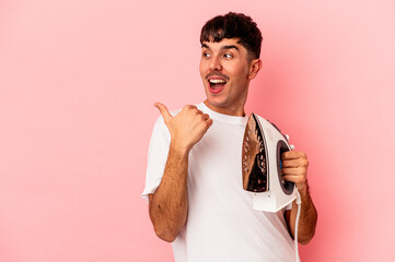 Wall Mural - Young mixed race man holding an iron isolated on pink background points with thumb finger away, laughing and carefree.