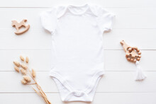 Mockup Of White Baby Bodysuit On Wood Background With Dried Flowers And Toys, Flat Lay. Blank Baby Clothes Template In Neutral Color.