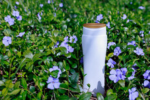 MockUp Bottle Of Shampoo Standing In A Clearing With Flowering Periwinkle. Purple Vinca Minor, Flowers In A Clearing With Bright Sleep And Sharp Shadows. White Unbranded Dispenser Bottle