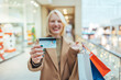 © Dragana Gordic - A happy beautiful blonde woman is smiling at her credit card, holding shopping bags, copying the space on the background of the shopping center.