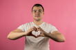© Регина Ерофеева - A young handsome man in a white T-shirt on an isolated pink background makes a heart shape with his hands. Concept: love, Valentine's Day, romance