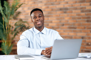 Portrait of confident successful smart handsome african american businessman, ceo or top manager, sitting in modern office, using laptop for work, wearing formal wear, looking at camera, smiling