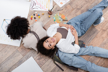 Top view of african american woman smiling at camera near boyfriend painting on floor in living room.