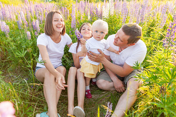 Wall Mural - Happy family mother father embracing kids outdoor. Woman man baby child and teenage girl sitting on summer field with blooming flowers background. Happy family mom dad and daughters playing on meadow