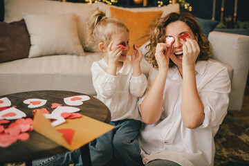 Mother and daughter making Valentine's day cards using color paper, scissors and pencil, sitting by the table in cozy room