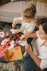 Mother and daughter making Valentine's day cards using color paper, scissors and pencil, sitting by the table in cozy room