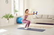 © Studio Romantic - Beautiful and slender young woman performs sports exercises at home using dumbbells. Smiling girl in comfortable sportswear sitting down slightly and stretching her arms forward holding two dumbbells.