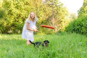 Wall Mural - A blonde young woman in a light dress throws a flying disc after which a cute dog runs