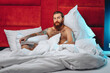 Attractive handsome young man with a bushy beard and tattoos on his arms and legs with a book in his hands smiling and sitting on a bed with gray linen and pillows 