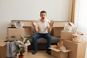 Portrait of angry man wearing white T-shirt and jeans sitting on sofa surrounded with cardboard boxes, raised arms, screaming with aggressive, moving and relocating.