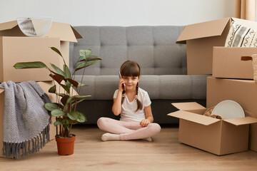 Portrait of smiling little girl with pigtails wearing casual style attire sitting on a floor near sofa and talking phone, boasting her moving in a new apartment with her family.