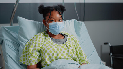 Wall Mural - Portrait of young patient with sickness wearing face mask in hospital ward. Sick woman in bed waiting on medical assistance against disease from doctor during coronavirus pandemic.