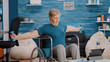 Aged woman stretching arms muscles with dumbbells, sitting in wheelchair and watching video of workout lesson on tablet. Senior person with physical disability using weights to exercise.