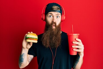 Canvas Print - Redhead man with long beard eating a tasty classic burger and drinking soda making fish face with mouth and squinting eyes, crazy and comical.