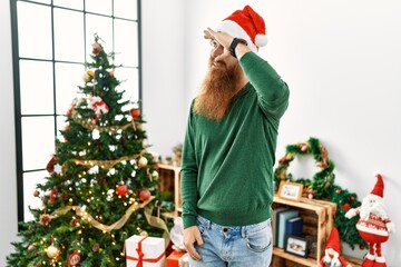 Poster - Redhead man with long beard wearing christmas hat by christmas tree very happy and smiling looking far away with hand over head. searching concept.