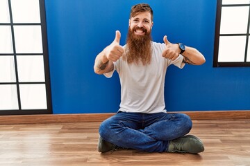 Sticker - Redhead man with long beard sitting on the floor at empty room approving doing positive gesture with hand, thumbs up smiling and happy for success. winner gesture.