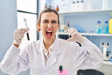 Canvas Print - Young woman working at scientist laboratory holding blood sample angry and mad screaming frustrated and furious, shouting with anger. rage and aggressive concept.