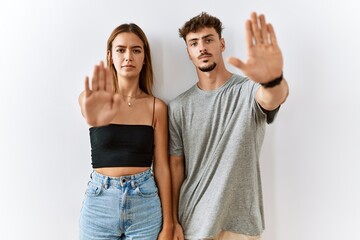 Wall Mural - Young beautiful couple standing together over isolated background doing stop sing with palm of the hand. warning expression with negative and serious gesture on the face.