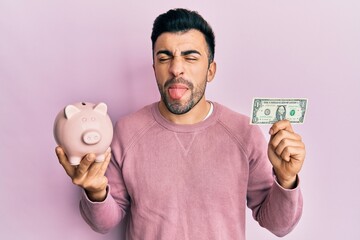 Young hispanic man holding one dollar banknote and piggy bank sticking tongue out happy with funny expression.