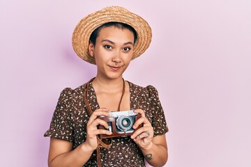 Poster - Beautiful hispanic woman with short hair wearing summer hat holding vintage camera relaxed with serious expression on face. simple and natural looking at the camera.