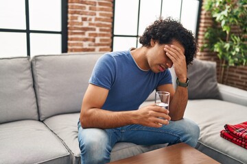 Sticker - Young hispanic man unwell drinking water sitting on sofa at home