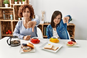 Sticker - Family of mother and down syndrome daughter sitting at home eating breakfast looking unhappy and angry showing rejection and negative with thumbs down gesture. bad expression.