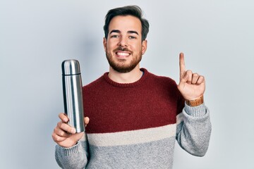 Handsome caucasian man with beard holding thermo smiling with an idea or question pointing finger with happy face, number one