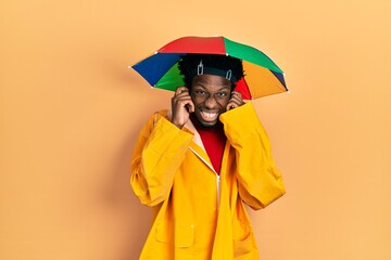 Poster - Young african american man wearing yellow raincoat covering ears with fingers with annoyed expression for the noise of loud music. deaf concept.