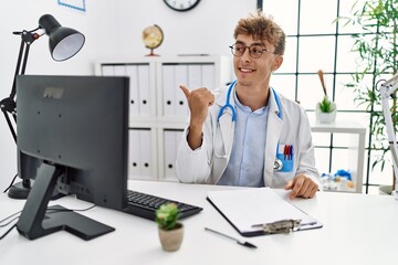 Wall Mural - Young caucasian doctor man working at the clinic smiling with happy face looking and pointing to the side with thumb up.