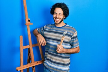 Poster - Handsome hispanic man standing by empty easel stand holding brushes winking looking at the camera with sexy expression, cheerful and happy face.