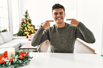 Sticker - Young hispanic man sitting on the table by christmas tree smiling cheerful showing and pointing with fingers teeth and mouth. dental health concept.