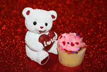 White Teddy Bear Holding Red Glitter Heart Sitting Beside A Cupcake, On A Red Glitter Background.