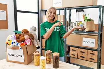 Poster - Young caucasian woman wearing volunteer t shirt at donations stand amazed and smiling to the camera while presenting with hand and pointing with finger.