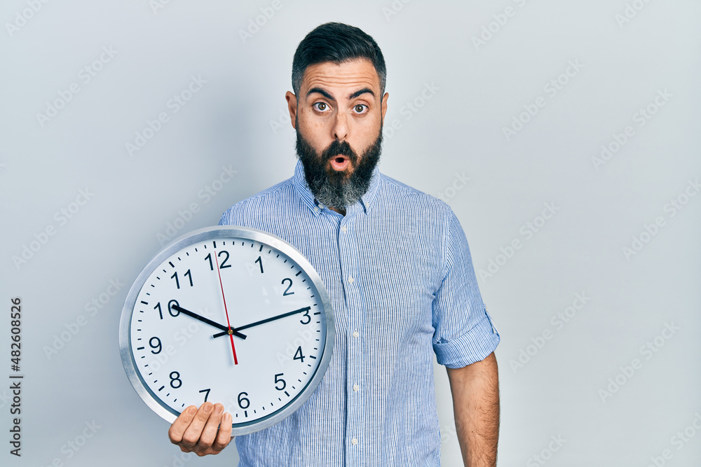 Young hispanic man holding big clock scared and amazed with open mouth for surprise, disbelief face