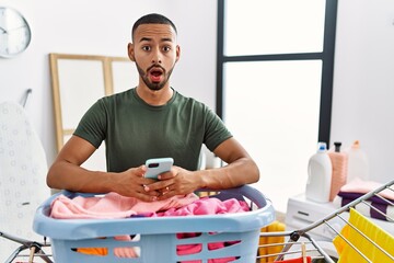 Poster - African american man doing laundry using smartphone in shock face, looking skeptical and sarcastic, surprised with open mouth