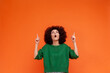 Portrait of shocked woman with Afro hairstyle wearing green casual style sweater looking and pointing up with fingers, presenting advertising area. Indoor studio shot isolated on orange background.
