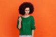 Portrait of serious woman with Afro hairstyle wearing green casual style sweater looking at camera, showing warning attention sign. Indoor studio shot isolated on orange background.