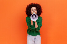 Attractive Woman With Afro Hairstyle Wearing Green Casual Style Sweater Screaming Loud Using Megaphone, Making Announcement. Presentation. Indoor Studio Shot Isolated On Orange Background.