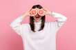 Positive woman covering eyes with two half slices of grapefruit, detox and healthy raw fresh food, wearing white casual style sweater. Indoor studio shot isolated on pink background.