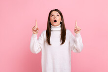Surprised Happy Brunette Girl Looking Astonished And Pointing Up Copy Space, Showing Empty Place, Wearing White Casual Style Sweater. Indoor Studio Shot Isolated On Pink Background.
