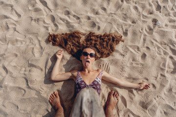 Wall Mural - Overhead view of a beautiful and playful girl in a bikini and sunglasses looking straight into the camera and sticking out her tongue while lying on the sand of the beach. Joy concept