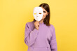 Woman with dark hair removing white mask from face showing his smiling expression, good mood, pretending to be another person, wearing purple hoodie. Indoor studio shot isolated on yellow background.