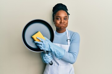 Poster - African american woman with braided hair wearing apron holding scourer washing pan puffing cheeks with funny face. mouth inflated with air, catching air.