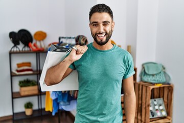 Canvas Print - Young handsome man with beard holding shopping bags at retail shop winking looking at the camera with sexy expression, cheerful and happy face.