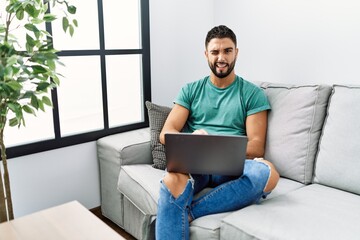 Canvas Print - Young handsome man with beard using computer laptop sitting on the sofa at home winking looking at the camera with sexy expression, cheerful and happy face.